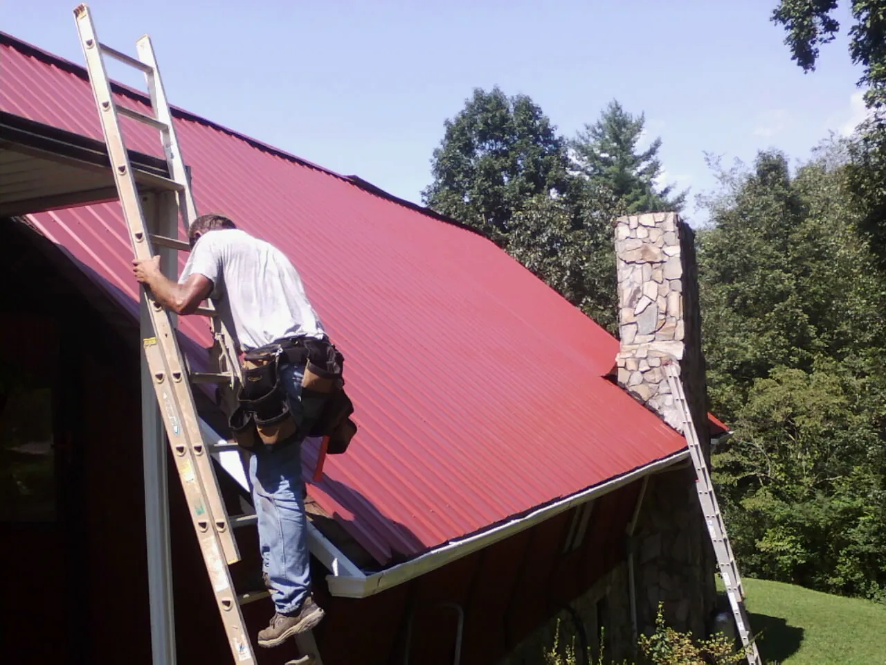 Roofer on a ladder beside a red metal roof and stone chimney — Soffit & Fascia Repair by Roofing Brothers in York Haven